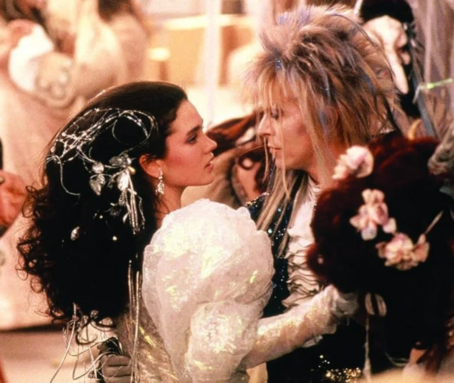A still from the 1986 film Labyrinth, starring David Bowie and Jennifer Connelly. Close-up of a woman and a man facing each other, dressed in elaborate, fantasy costumes and wigs.