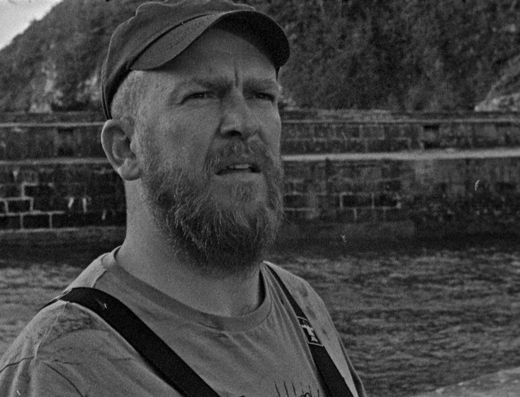 A man with a beard wearing a cap and a t-shirt standing outdoors with a brick wall and hills in the background. This is a scene from the 2019 film Bait Directed by Mark Jenkin and starring Edward Rowe