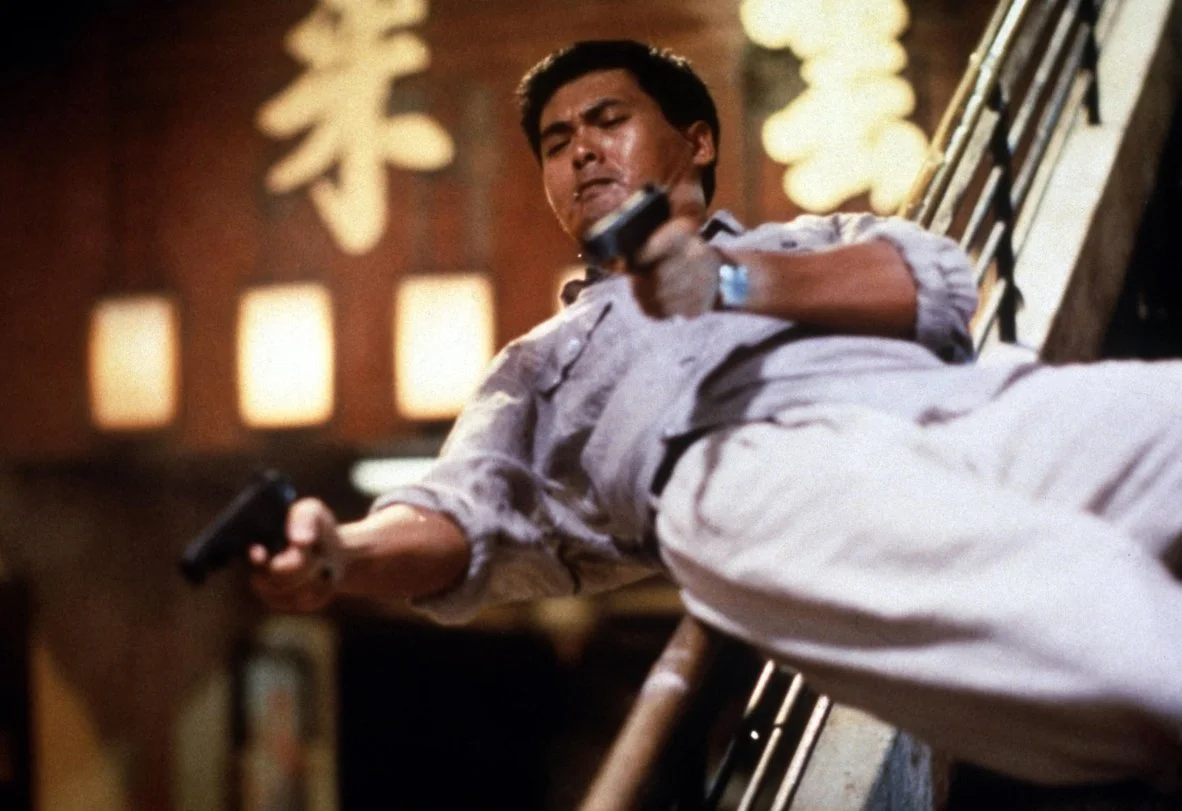 A man sliding down the banister of a set stairs holding a gun in each hand, he is dressed in white shirt and trousers, he looks awesome. This is a still from John Woo's 1992 film Hard Boiled starring Chow Yun-fat.