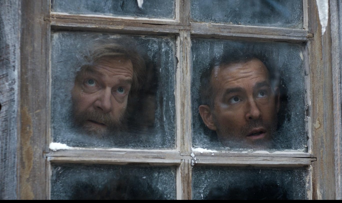 Two men looking through a dirty window with four panes, one with a beard and the other clean-shaven. Mayday (2026) starring Kenneth Branagh and Ryan Reynolds. Directed by John Francis Daley and Jonathan Goldstein.
