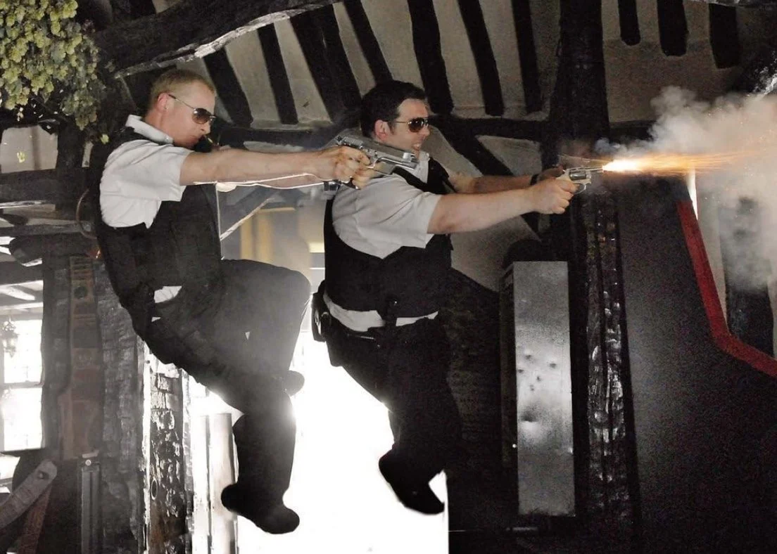 Two men dressed as police officers diving threw the air shooting guns inside of a pub. This is a still from the 2007 film Hot Fuzz Directed by Edgar wright and starring Nick Frost and Simon Pegg.