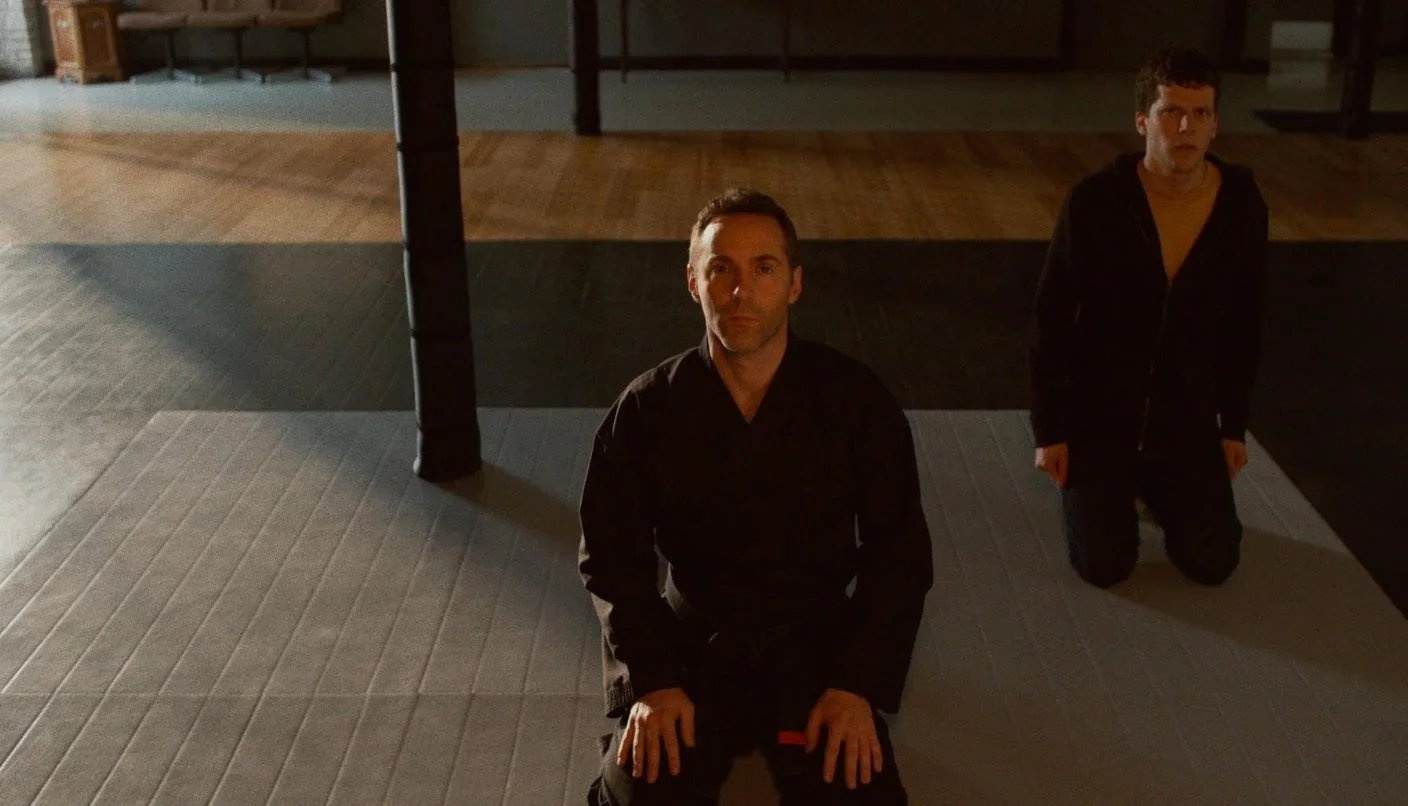 Two men are kneeling on the floor in a dimly lit room, looking up at the camera. A man is wearing a black gi the other man is wearing a black hoodie with a yellow t-shirt. 2019 Film The Art of Self-Defense directed by Riley Stearns.