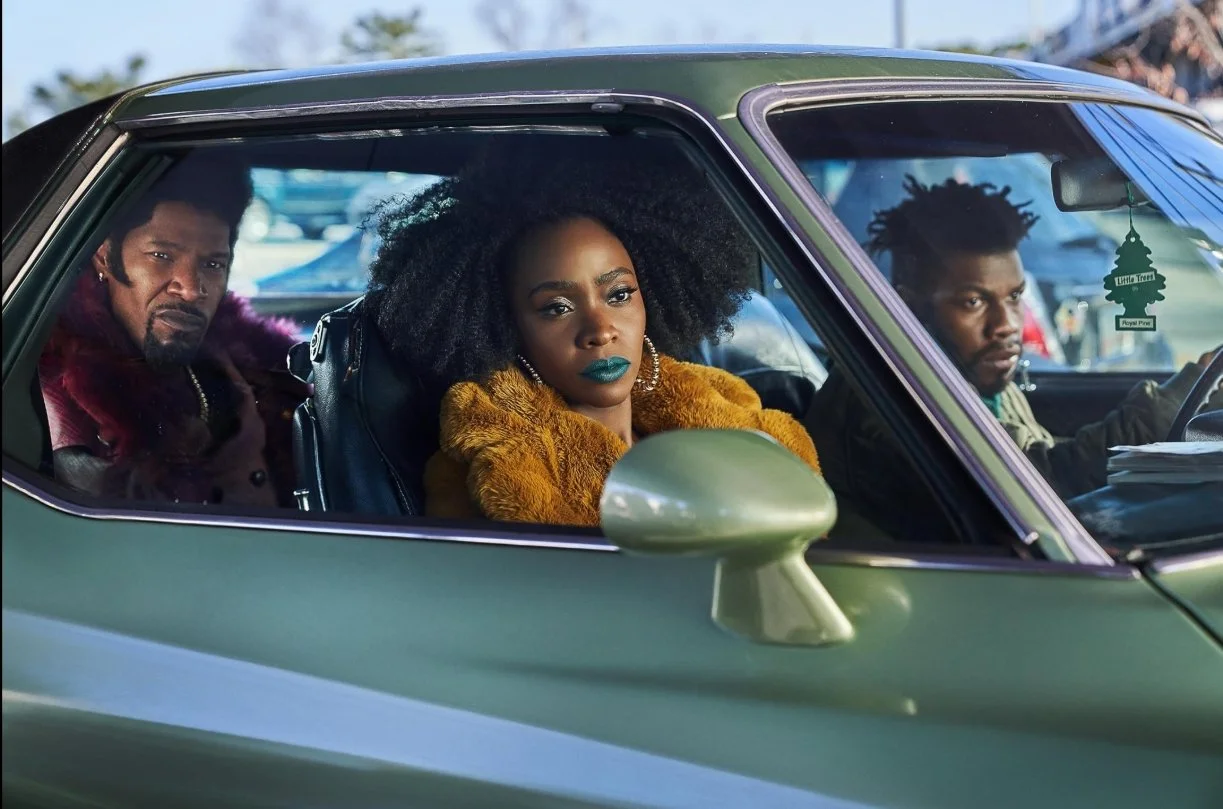 A woman with has curly hair, teal lipstick wearing a mustard-coloured coat sits in a green car with two men, one has a beard, short hair, and is wearing a purple coat, the other man on the right has short hair. They Cloned Tyrone (2023).