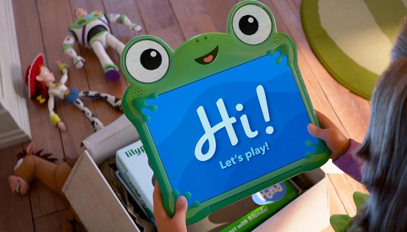 Child holding a tablet with a frog design screen that says 'Hi! Let's play!'. Toys including Woody from Toy Story, Buzz Lightyear, and Jessie are on the wooden floor nearby. From Toy Story 5 (2026) Starring, Joan Cusack, Tom Hanks, Tim Allen.