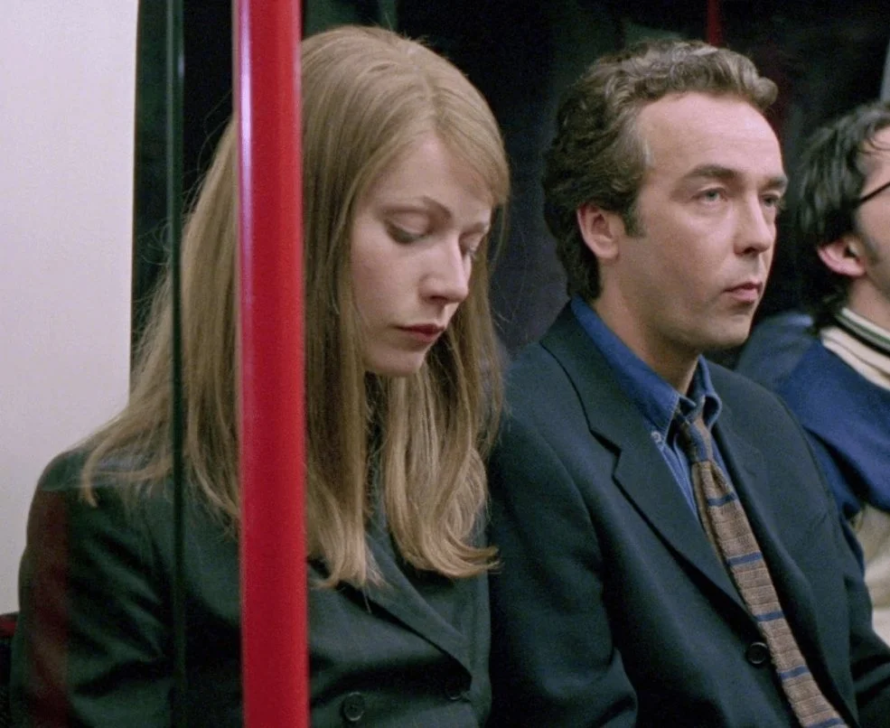 A film still from the rom-com film Sliding Doors (1998) featuring Gwyneth Paltrow and John Hannah. A man and woman sit next to each other on a train.