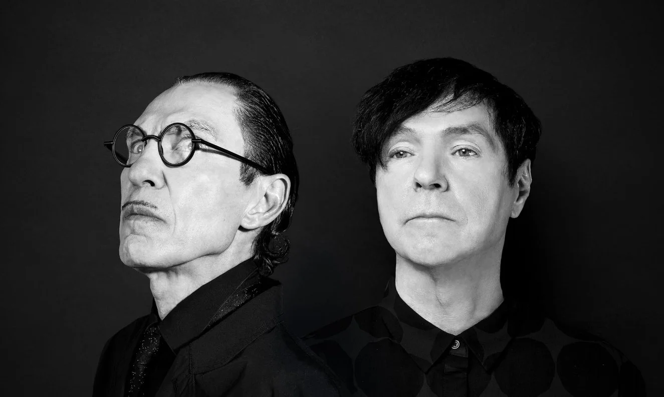 Black and white photo of two men, one on the left wearing glasses and with slicked-back hair, and the other on the right with dark, tousled hair, both looking serious against a dark background. Sparks, Ron and Russell Mael.