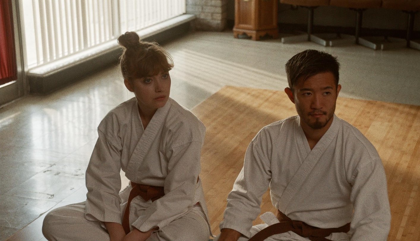 A young woman and a young man in martial arts uniform sitting on the floor in a dojo, with sunlight coming through the windows behind them. This is a scene from the 2019 film The Art of Self-Defense, directed by Riley Stearns Starring Jesse Eisenberg