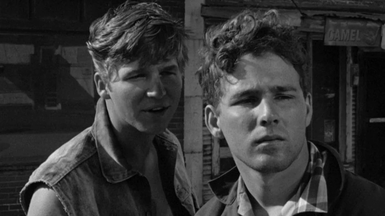 A black and white film still from The Last Picture Show (1971) starring Jeff Bridges and Timothy Bottoms, they are standing together talking while one looks off into the distance.
