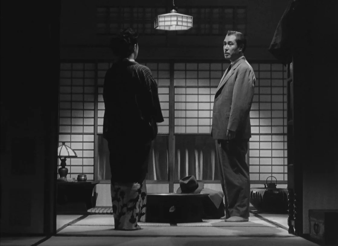 A black and white photo of two people facing each other in a traditional Japanese room. One is a woman in a kimono the other is a man in a suit. They are standing on tatami mats next to a low table. Ozu's The Flavour of Green Tea Over Rice.