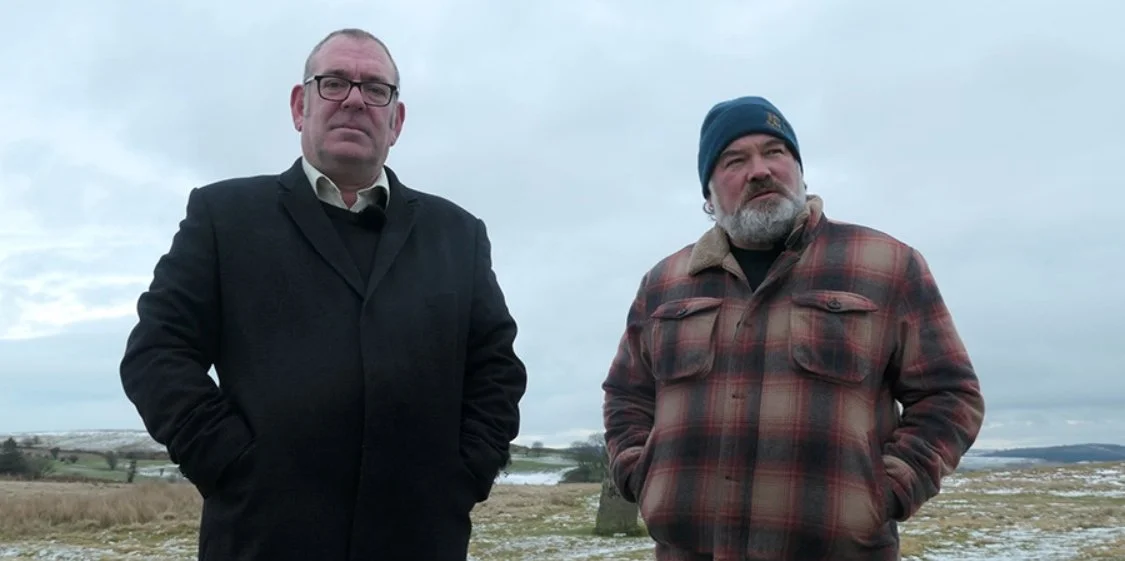 Two men standing outdoors in a cloudy landscape, one wearing a black coat and glasses, the other in a plaid jacket and beanie. Musician Robert Lloyd and Comedian Stewart Lee in the 2020 Documentary film King Rocker.