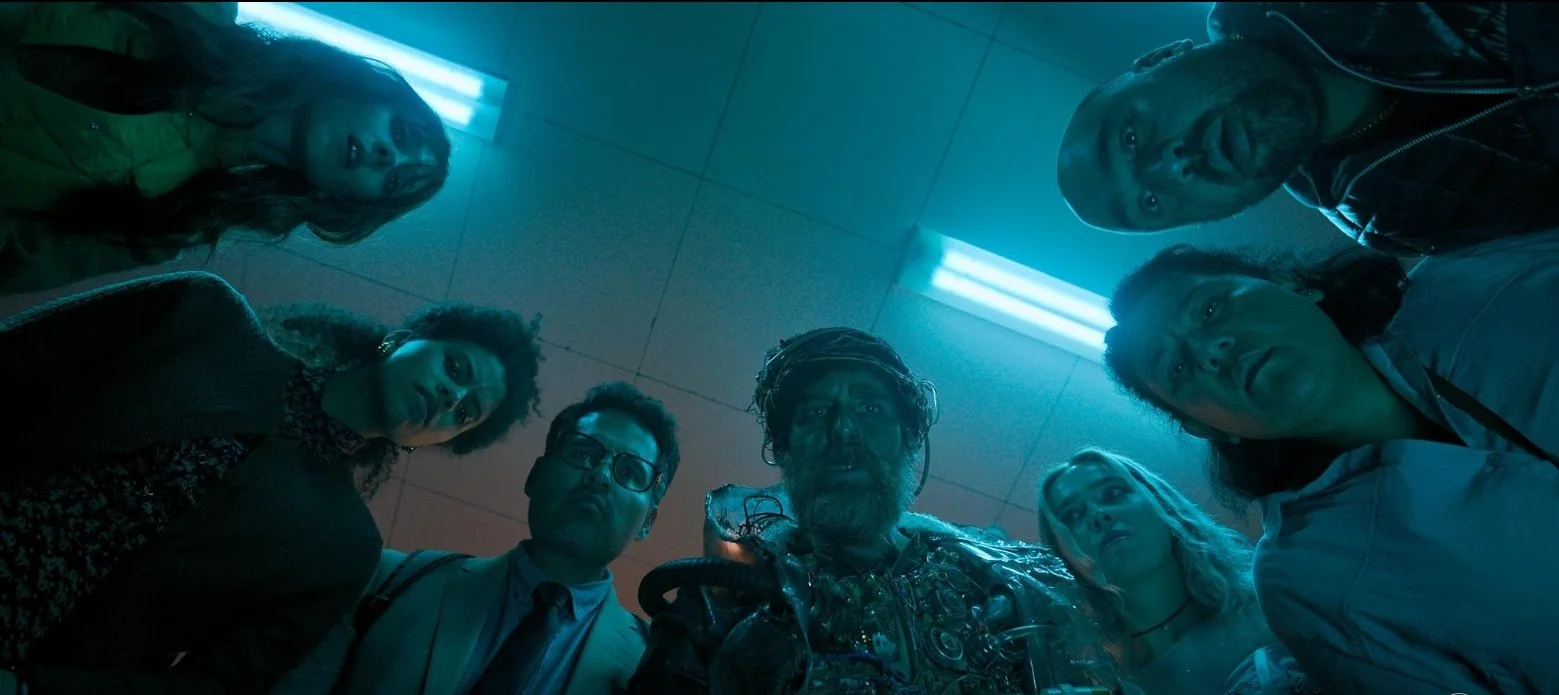 A group of seven people, including a bearded man in elaborate armor, peer down at the camera under blue fluorescent lighting. Good Luck, Have Fun, Don't Die (2026) directed by Gore Verbinski and starring Sam Rockwell, Juno Temple and Haley Lu Richard