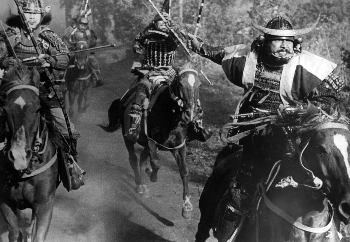 A group of Japanese warriors on horseback, dressed in traditional attire, riding through a forested area. This is a scene from the 1957 film Throne of Blood directed by Akira Kurosawa and starring Toshiro Mifune and Isuza Yamada.