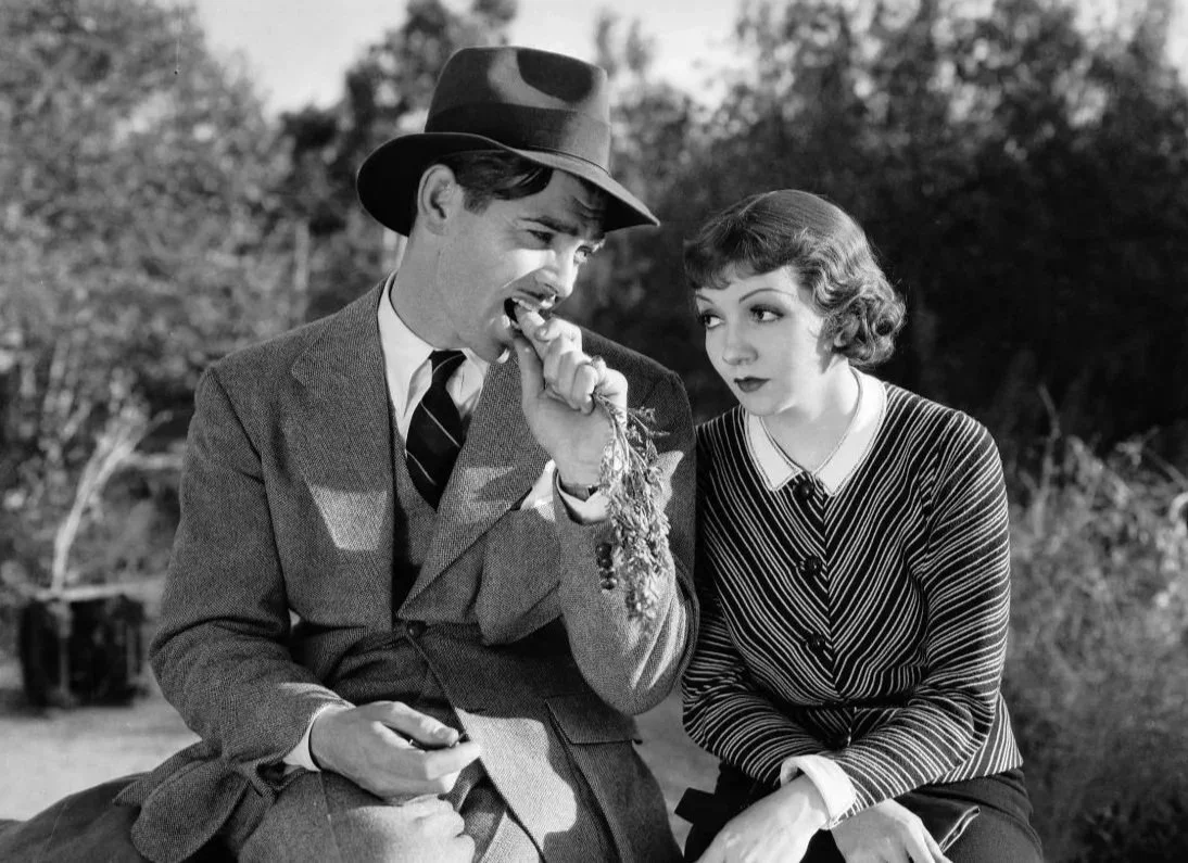 A black and white still from the rom-com film, It Happened One Night (1934) featuring Clark Gable, Claudette Colbert. a couple sit together, the man has a hat and is eating a carrot.