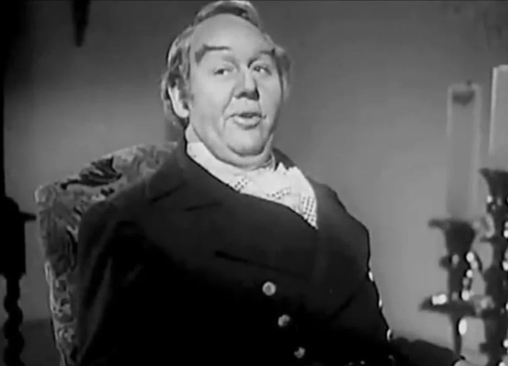 A black and white still from the film Jamaica Inn (1939) starring Charles Laughton, here he is sitting looking pompous while wearing a suit and cravat.