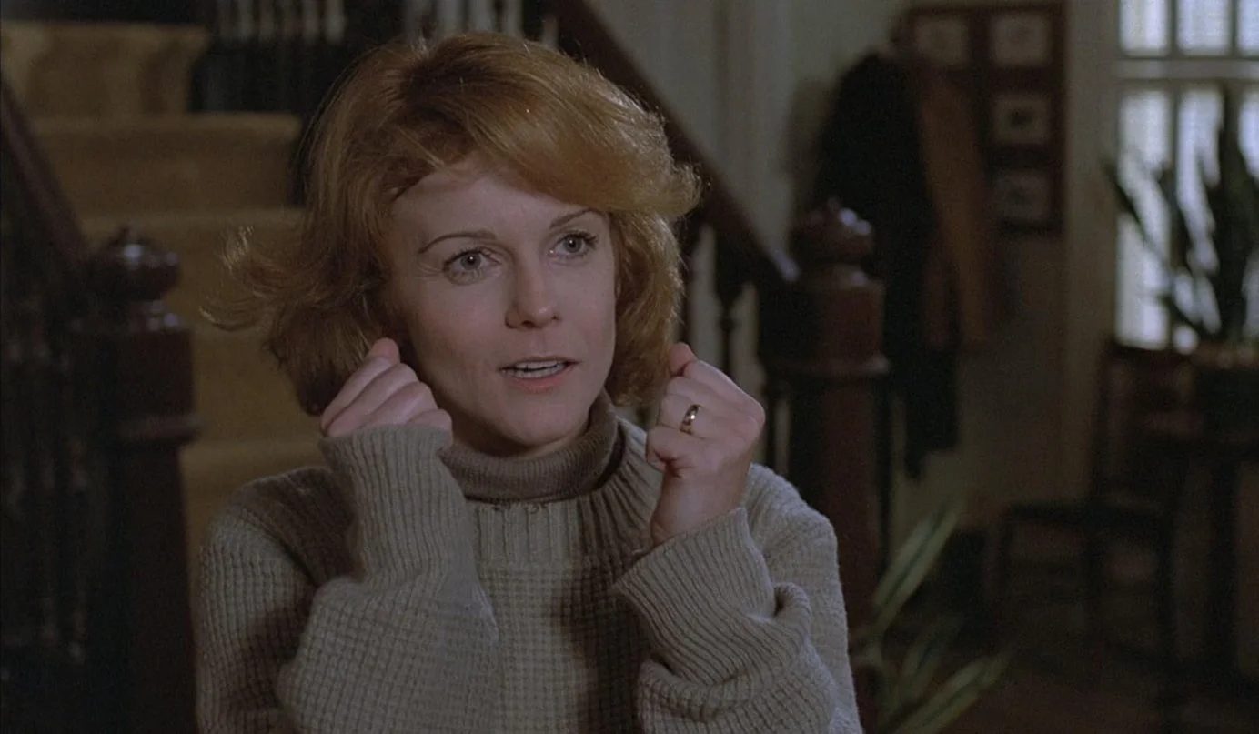 A woman with red hair wearing a beige turtleneck sweater, holding her fists near her face, in a warmly lit room with wooden stairs and framed pictures in the background. Magic (1978) starring Ann-Margret and Anthony Hopkins.