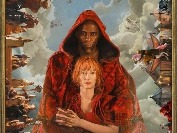 A film still from Three Thousand Years of Longing starring Tilda Swinton and Idris Elba, a djinn wearing a red hooded cape wraps his arms around a woman with ginger hair. the background is sky with fantastical skylines.