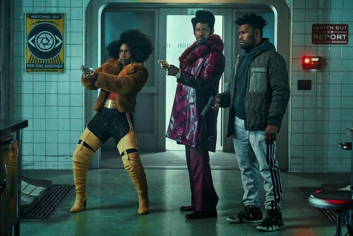 Three people with guns inside a room with tiled walls and warning signs and an open lift door behind them. They Cloned Tyrone (2023) directed by Juel Taylor starring Jamie Foxx, John Boyega, Teyonah Parris.