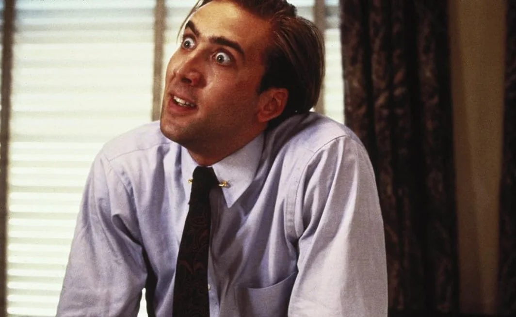 A man with an exaggerated facial expression, wide eyes, and raised eyebrows, wearing a light blue dress shirt and a dark tie, sitting in an office. This is a still from the 1988 film Vampire's Kiss starring Nicolas Cage.