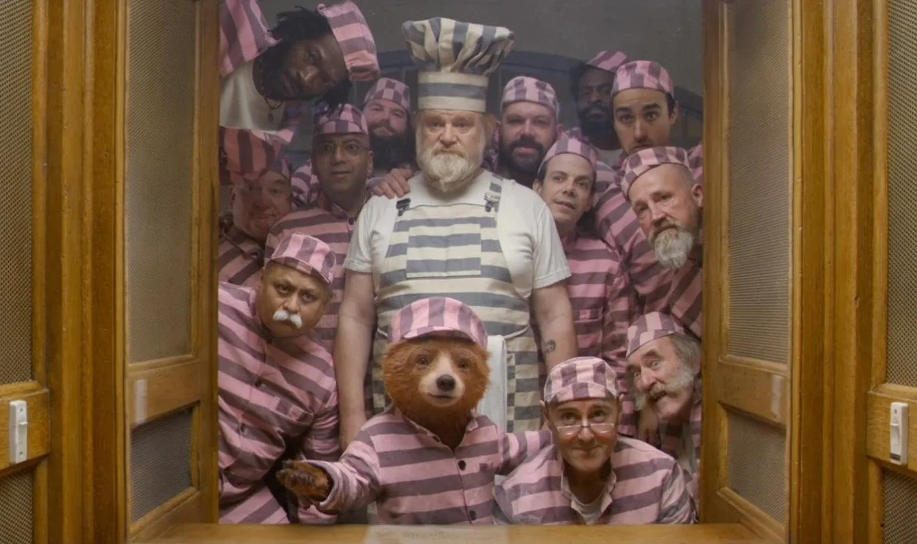 Scene from the 2017 film Paddington 2 starring Ben Whishaw and Brendan Gleeson. An image of a group of prisoners wearing pink and grey outfits, one of them is Paddington Bear, they are all looking towards the camera.