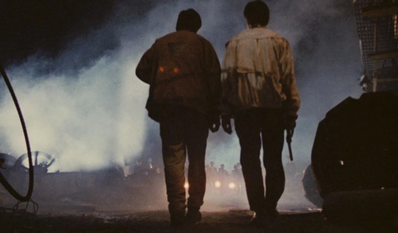 Two people walking in a smoky, dark area with burning debris and wreckage around them. This a still from the John Woo's 1986 action film A Better Tomorrow, starring Chow Yun-fat and Ti Lung.