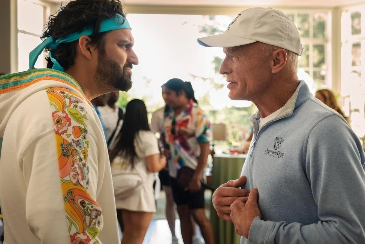 Two men face each other closely indoors during daytime, one wearing a colorful jacket and a blue headband, the other in a light gray cap and sweatshirt, This is a scene from the 2026 Apple Tv film The Dink, Starring Jake Johnson, Mary Steenbergen.