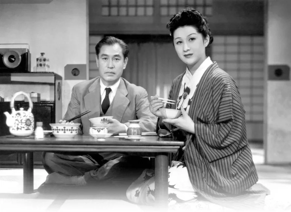 A black and white photo of a man and woman in traditional Japanese clothing, sitting at a low table with bowls and cups, in a traditional Japanese room. This is a scene from Ozu's The Flavour of Green Tea Over Rice (1952).