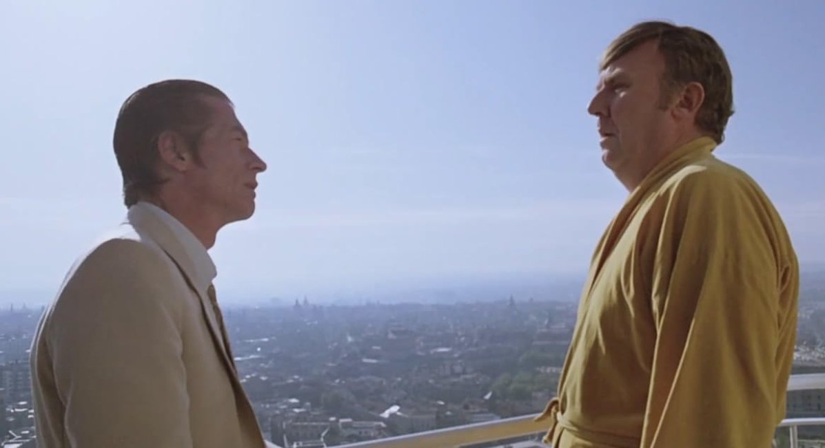 A film still from the crime gangster drama film The Hit (1984) featuring John Hurt and Bill Hunter. Two men are facing each other one in a cream suit the other a yellow bath robe.