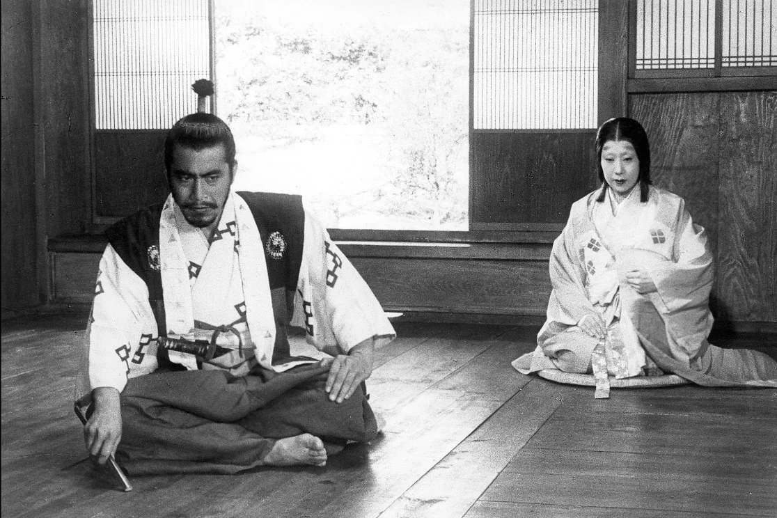 Two Japanese actors in traditional clothing sitting on the floor of a wooden room, with shoji screens and a window in the background. This is a still from the Akira Kurosawa film Throne of Blood (1957) starring Toshiro Mifune and Isuza Yamada.