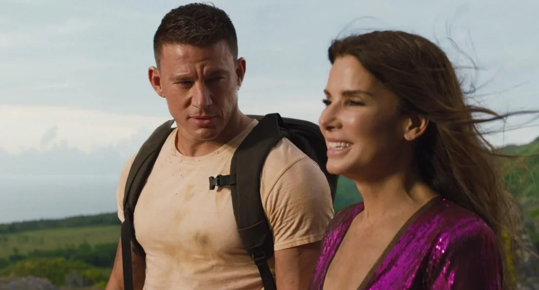 A film Still from the rom-com The Lost City (2022) featuring Channing Tatum and Sandra Bullock. A man wearing a backpack looks at a smiling woman wearing a glittery outfit.