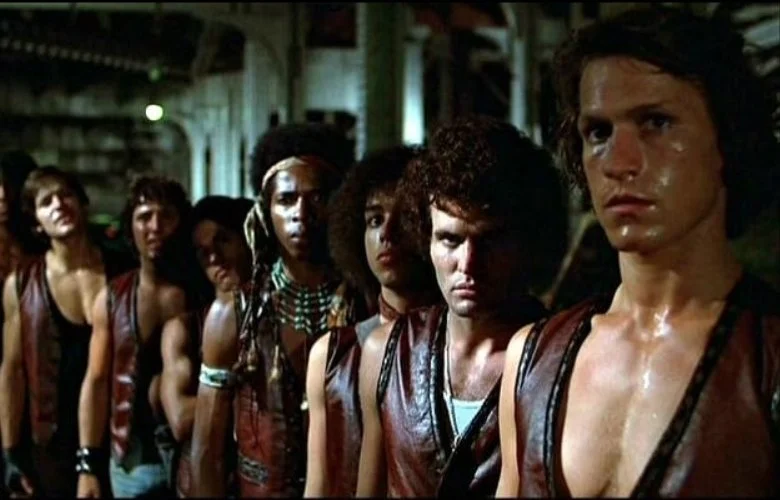 A gang of seven people standing in a line, wearing matching sleeveless leather vests, in a dimly lit setting. This is a still from the 1979 Walter Hill action film The Warriors, starring Michael Beck, James Remar and David Patrick Kelly.