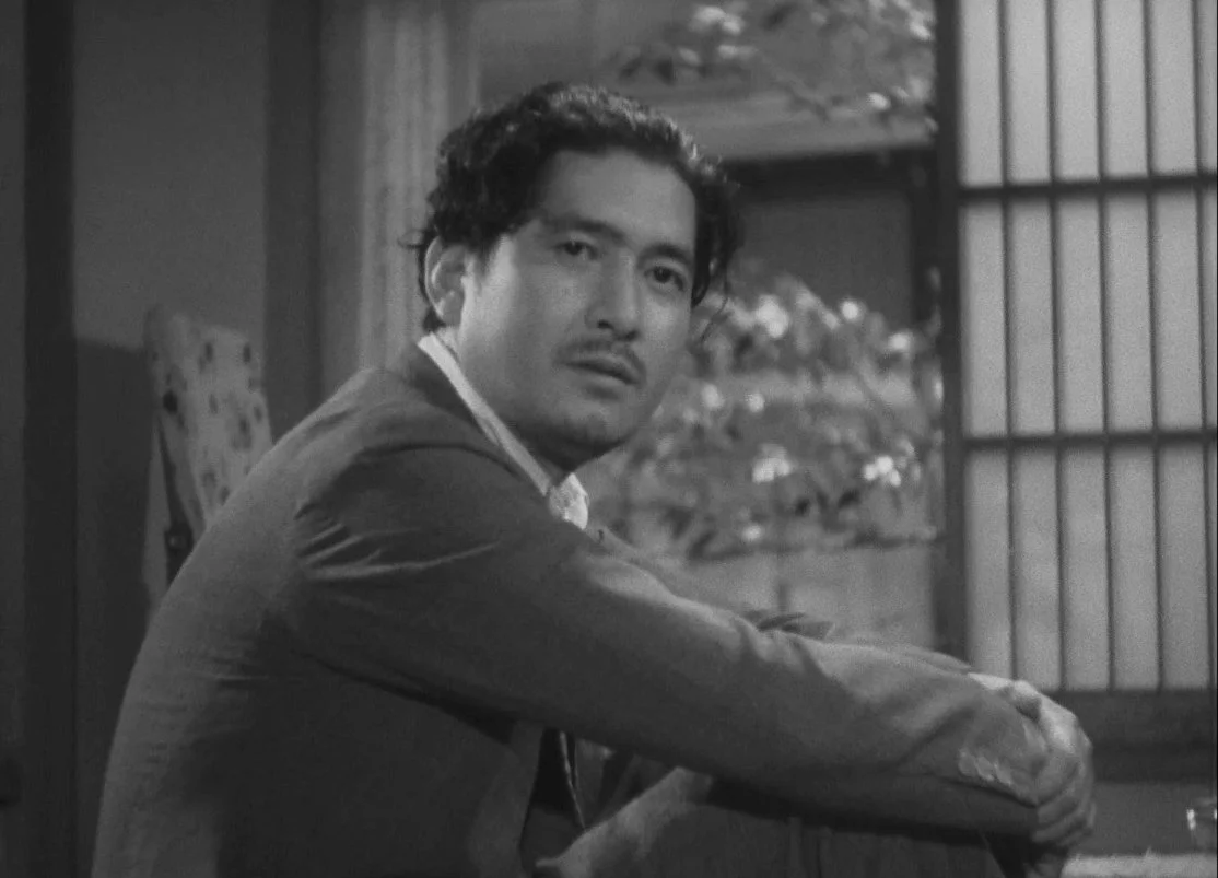 A black and white image of a man with wavy black hair, sitting indoors, looking towards the camera. Background features a paper screen and some foliage. this is a scene from Yasujirō Ozu 1948 Japanese film A Hen in the Wind