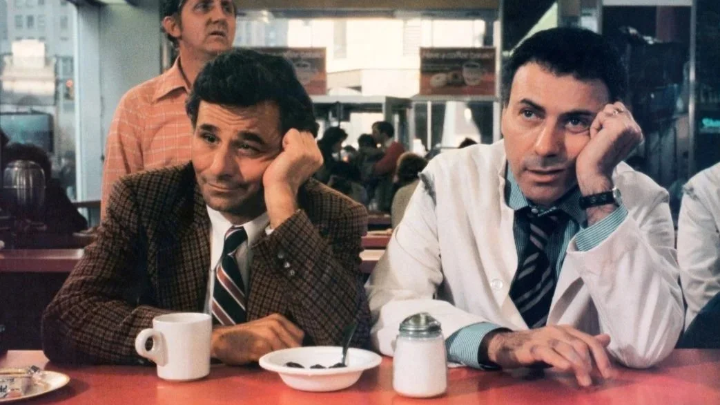 A film still from the comedy adventure film The In-Laws (1979) featuring Peter Falk and Alan Arkin sitting in a cafe wresting their heads on their left hand looking frustrated..