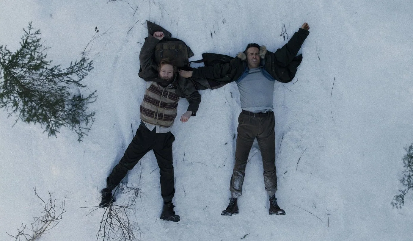 Two men are lying on snow-covered ground, handcuffed. This is a shot from the 2026 Apple TV film Mayday, starring Kenneth Branagh and Ryan Reynolds, written and directed by John Francis Daley and Jonathan Goldstein.