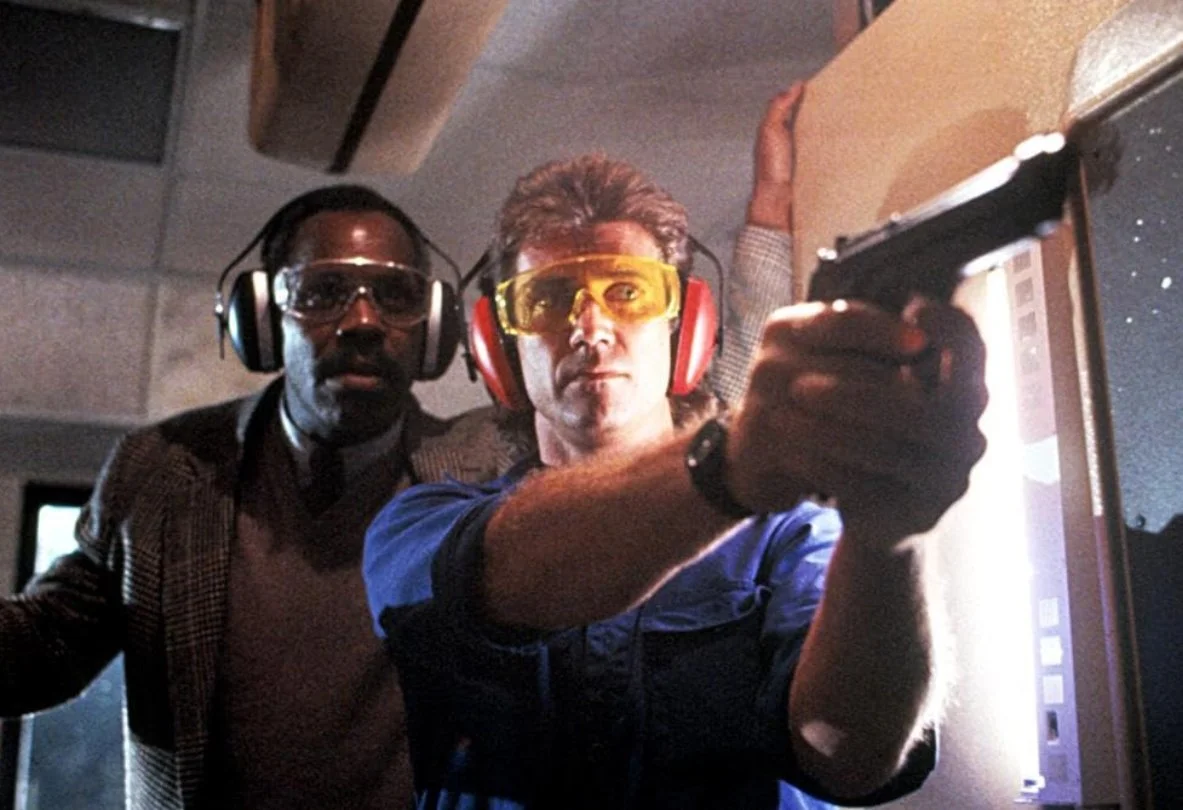 Two men in ear defenders and safety glasses aiming a gun in a shooting range. This is a still from Richard Donner's 1987 action film Lethal Weapon, starring Danny Glover and Mel Gibson.