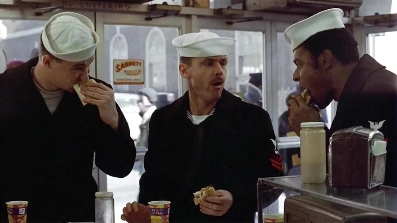 Three sailors in uniform eating sandwiches inside a diner. Two sailors are actively eating, one in the middle is engaged in conversation. This is a scene from The Last Detail with Jack Nicolson, Randy Quaid and Otis Young.