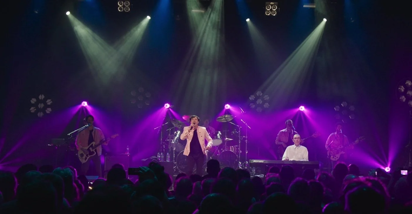 The band Sparks perform on stage under colorful purple light, the is singer in a beige jacket and a keyboard player in a white shirt in the foreground, with guitarists and a drummer in the background, in front of an audience.