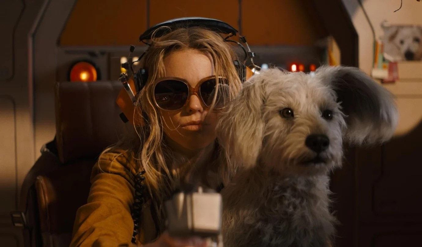 A still from the 2026 film Supergirl, starring Milly Alcock. In her spaceship, Supergirl is wearing sunglasses and head phones, while sitting with her dog Krypto on her knee. Krypto is a white fluffy dog with big ears.