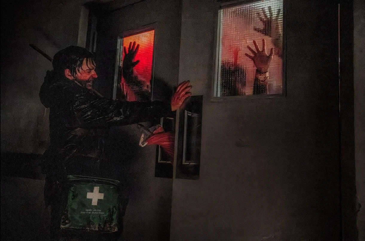 A person in a black coat with wet hair is holding a door shut with zombies or infected on the other side of the do. There are two windows with hands pressed against the glass from the inside, illuminated with red light. Resident Evil 2026 Cregger.