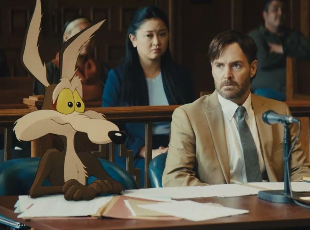 A courtroom scene showing a man in a tan suit and a woman with long dark hair behind him, with cartoon character Wile E. Coyote, this is a scene from the 2026 film coyote vs acme starring Eric Bauza, Will Forte John Cena.
