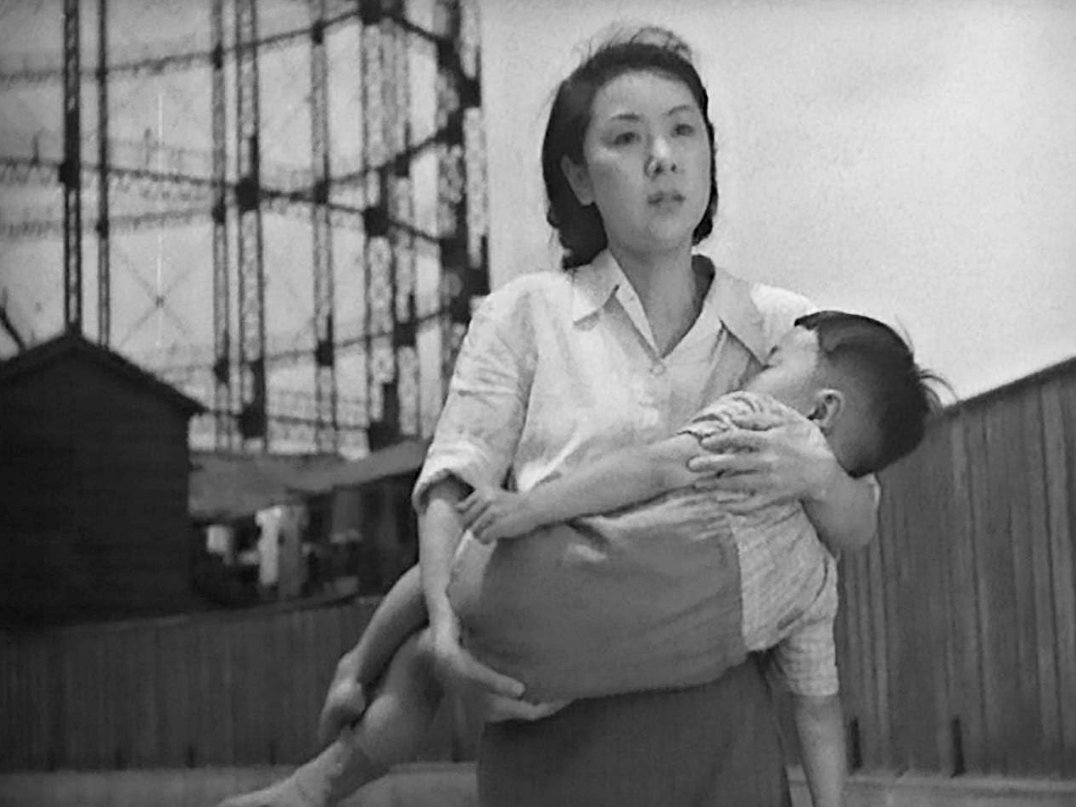 A woman carrying a young child in her arms, walking outdoors with a fence and industrial structures in the background, all in black and white. this is a scene from Yasujirō Ozu 1948 Japanese film A Hen in the Wind