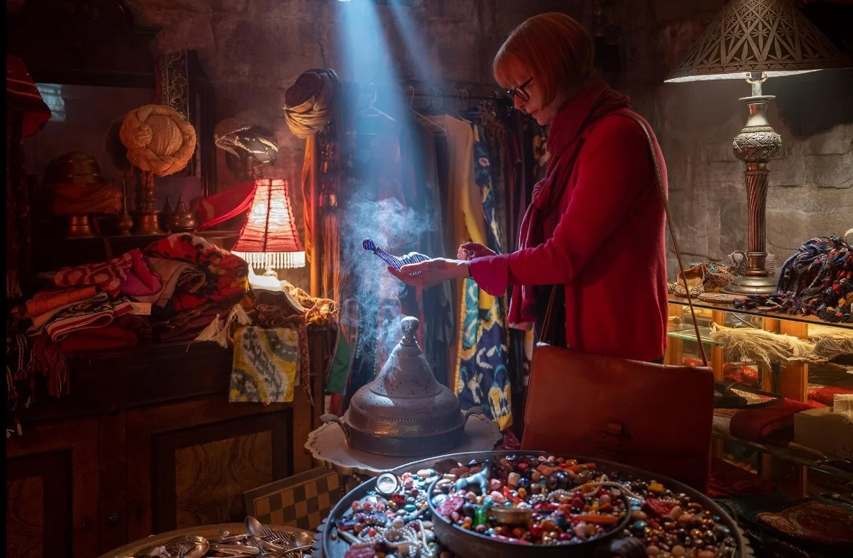 A still from the film Three Thousand Years of Longing (2022) with Tilda Swinton in a bazaar looking at curiosities and holding a strange, small, blue glass bottle with a sunbeam  highlighting it in the darkness.