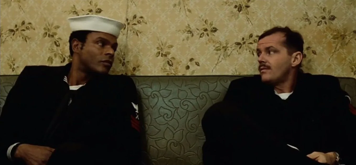 Two men in navy uniforms sitting on a bench with a yellow floral wallpaper background, having a conversation. This is a still from the 1973 film The Last Detail directed by Hal Ashby, written by Robert Towne starring Jack Nicolson and Randy Quaid.
