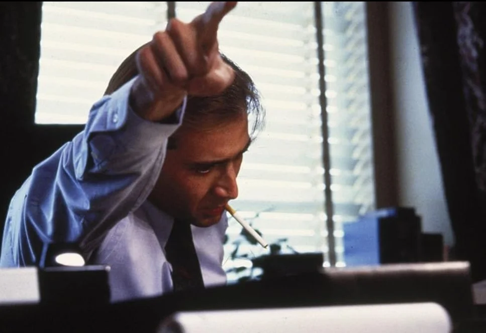 Man in office with white shirt and black tie, smoking cigarette, pointing his hand, sitting at desk with blinds in background. This is a scene from the 1988 film Vampire's Kiss Starring Nicolas Cage.