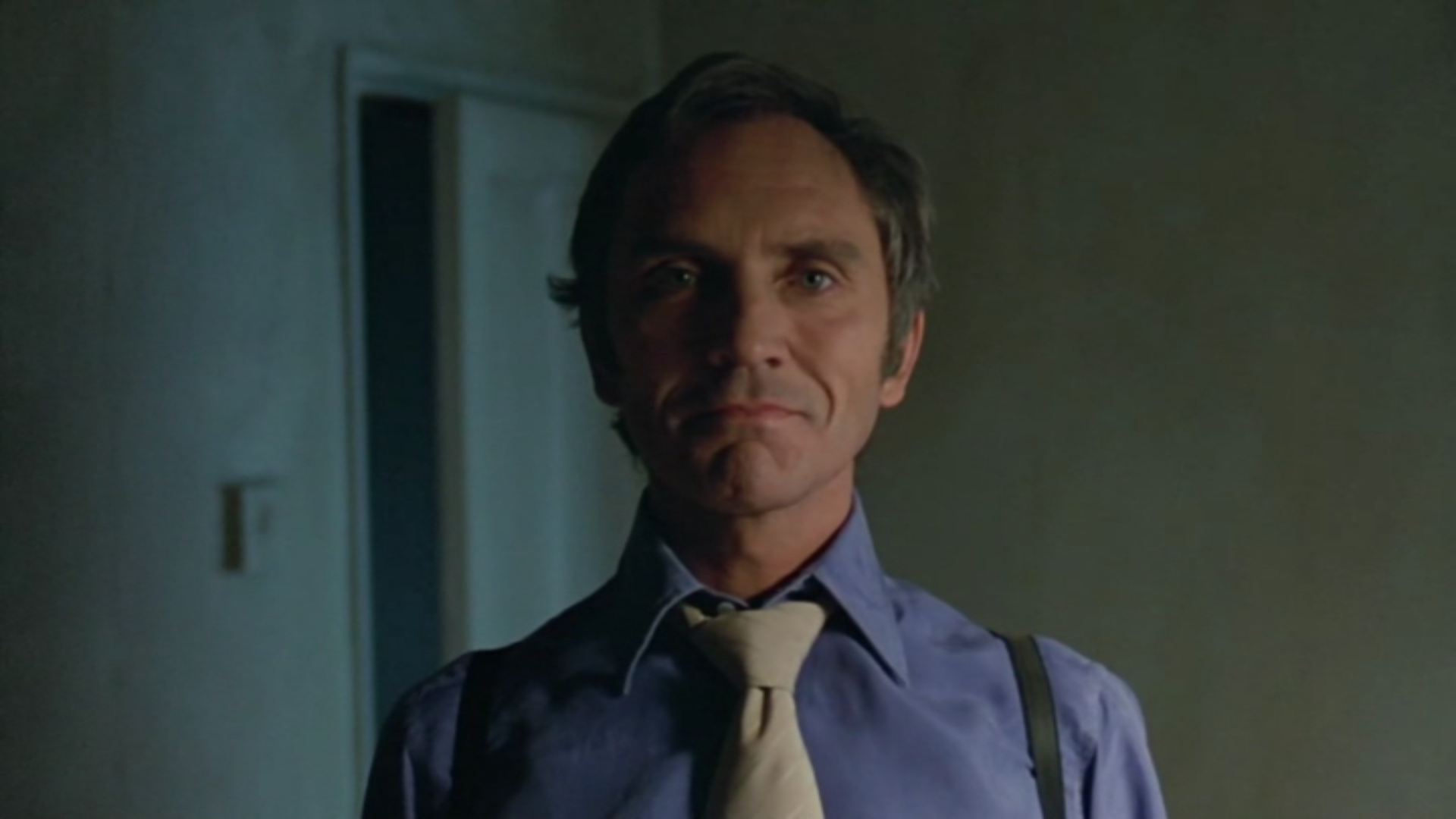 Terrance Stamp with a slight smile, wearing a blue shirt, tie, and suspenders, standing in a dimly lit room, in a still from Stephen Frears 1984 film The Hit.