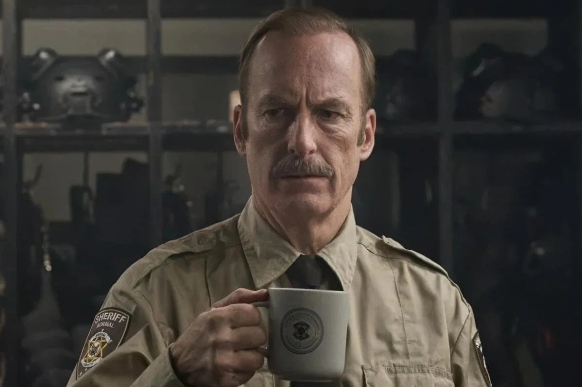 A middle-aged sheriff in uniform drinking from a mug with a police emblem on it. This is a still from the film Normal (2025), starring Bob Odenkirk, in Ben Wheatley's action film.