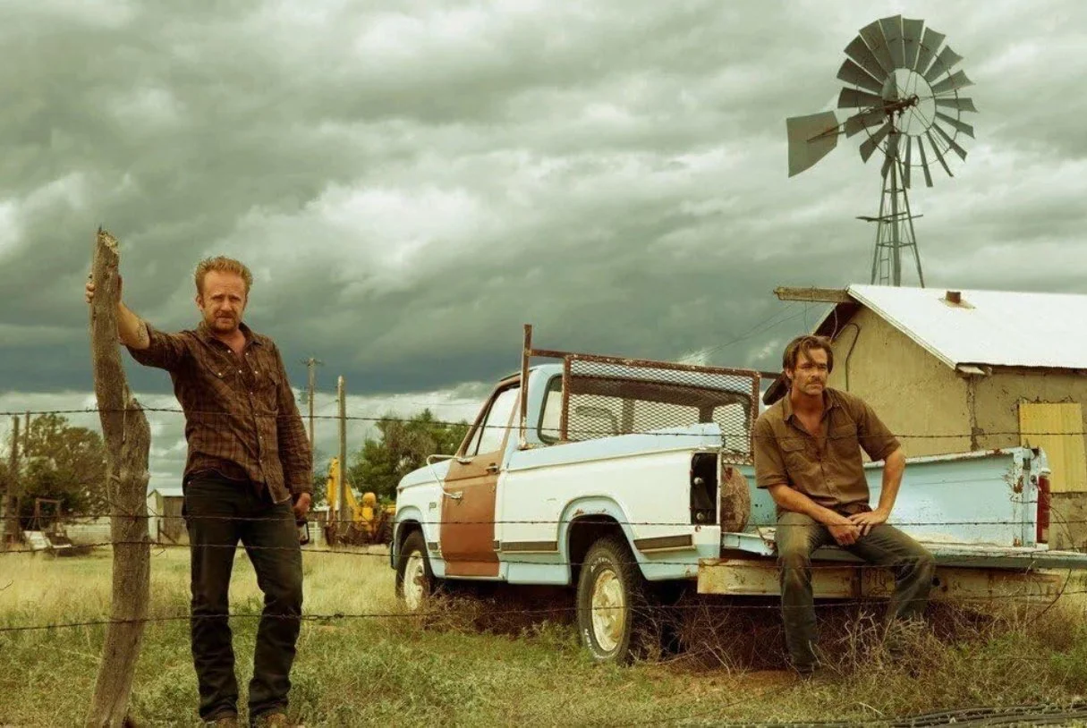 Two men in casual clothes in a rural setting, one leaning on a fence post and the other sitting on the tailgate of a weathered pickup truck, with a windmill and a cloudy sky in the background. Still from the film Hell or High Water.