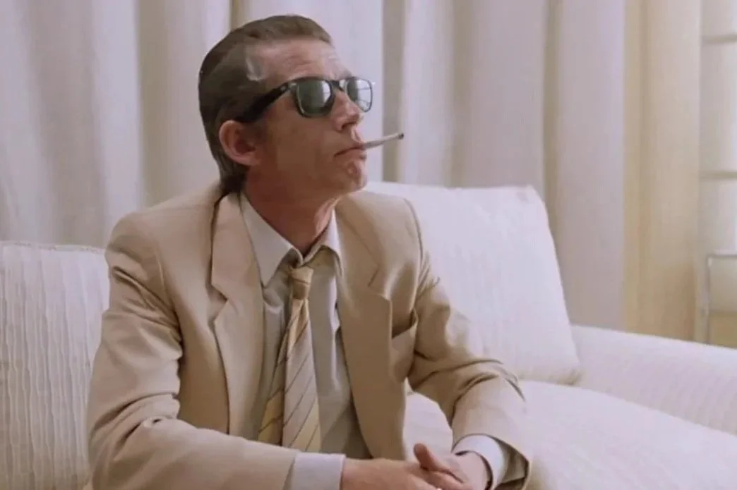 A film still from the crime gangster drama film The Hit (1984) Featuring John Hurt. A man is sitting on a white sofa, wearing a cream suit and sunglasses, while smoking.