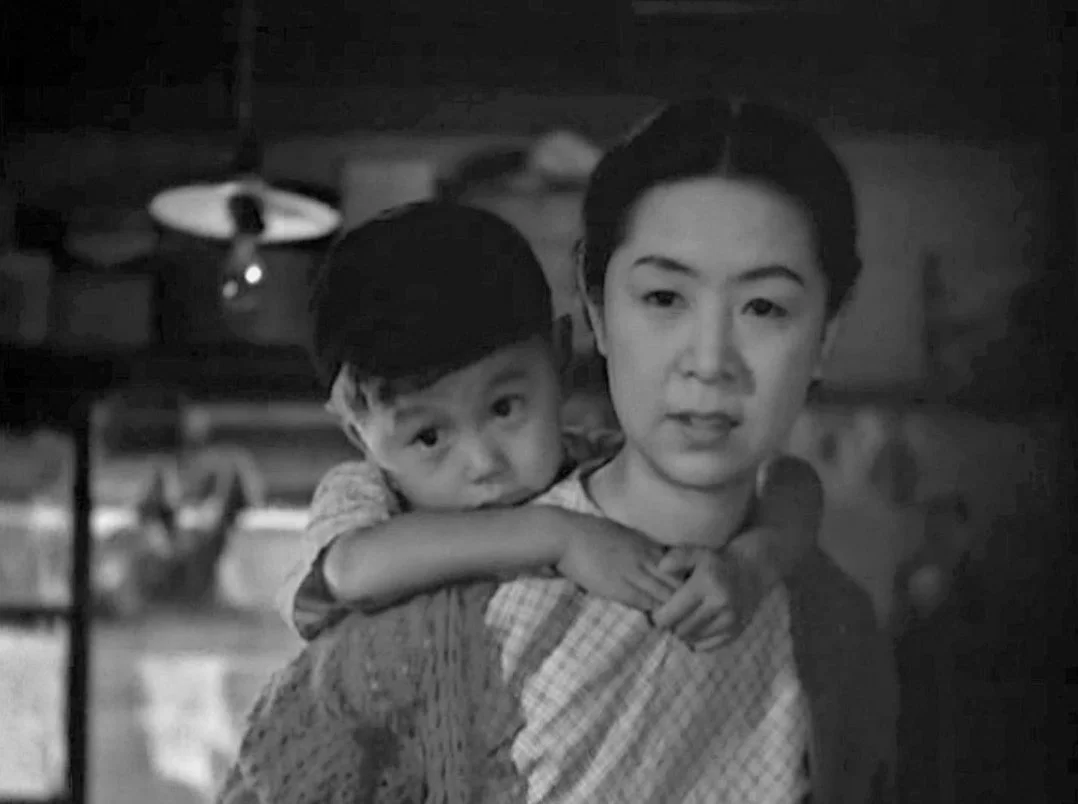 A woman with dark hair and a young boy with dark hair, carrying him on her shoulder in an indoor setting. This is a scene from Yasujirō Ozu 1948 Japanese film A Hen in the Wind. the image is black and white