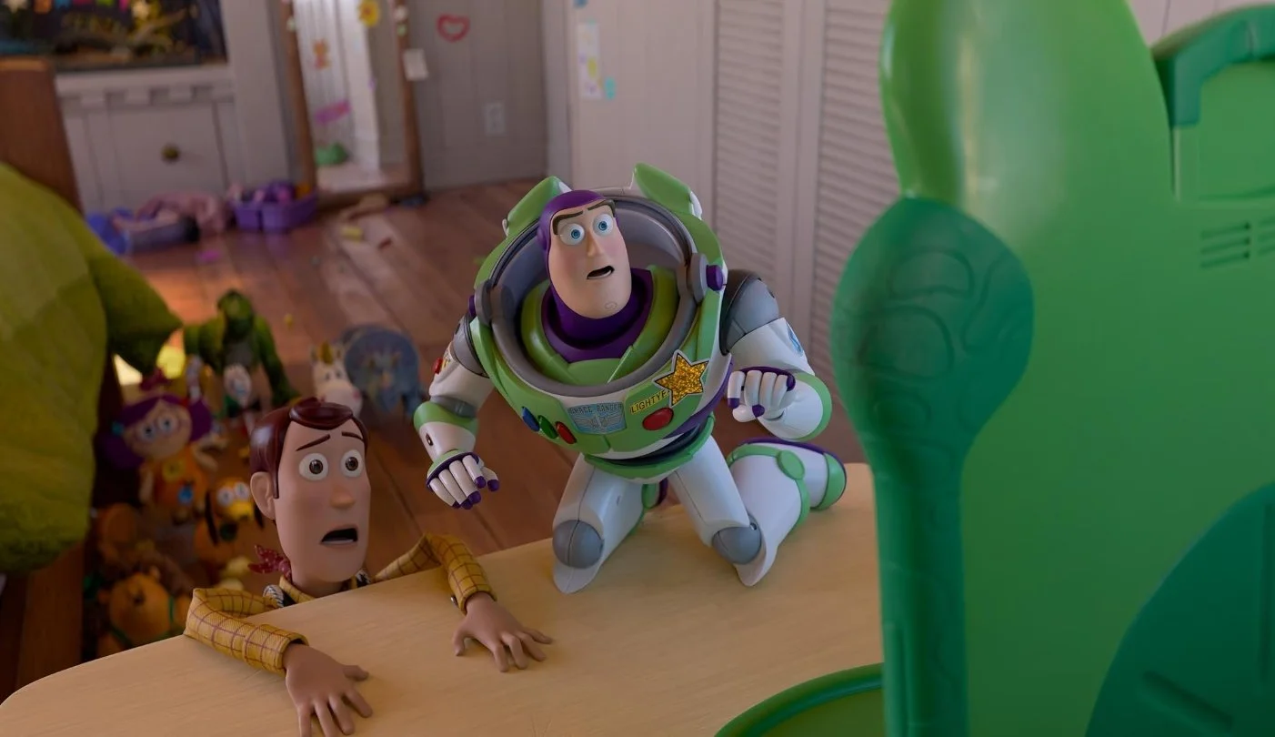 Buzz Lightyear and Sheriff Woody from Toy Story 5 (2026) looking startled at a green toy robot on a table surrounded by other toys. Starring Tom Hanks, Great Lee, Tim Allen.