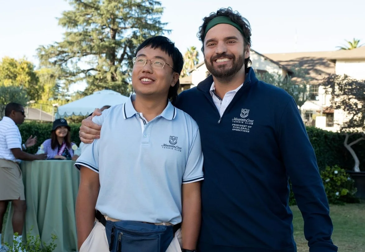 Two men standing outdoors, smiling, with one having his arm around the other's shoulder. The man on the left is wearing glasses and a light blue polo shirt, and the man on the right has dark curly hair, a beard, and is wearing a navy jacket. The Dink